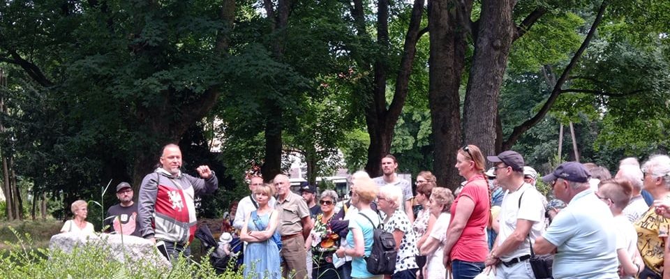 Grupa uczestników spaceru stoi w parku i słucha przewodnika, Tomasza Wieczorka