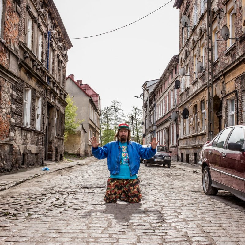 Kolorowe zdjęcie, mężczyzna klęczący na bruku, po obu stronach zaniedbane kamienice