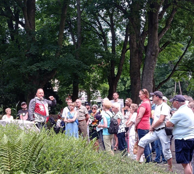 Grupa uczestników spaceru stoi w parku i słucha przewodnika, Tomasza Wieczorka